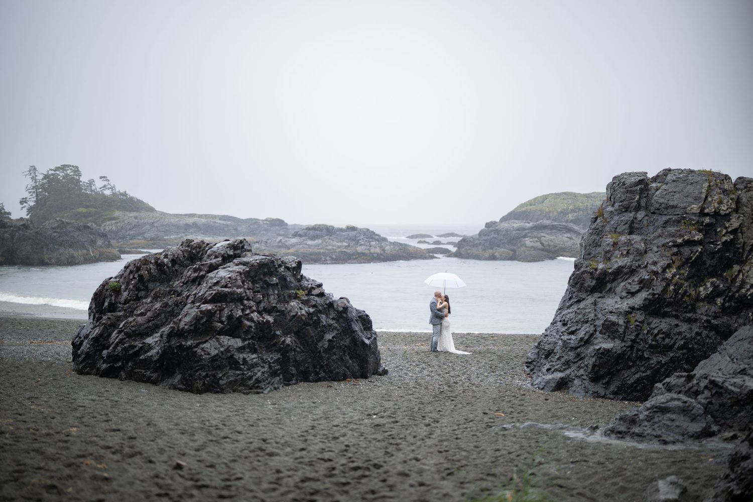 A distant view of a couple between large rock formations on a misty beach.