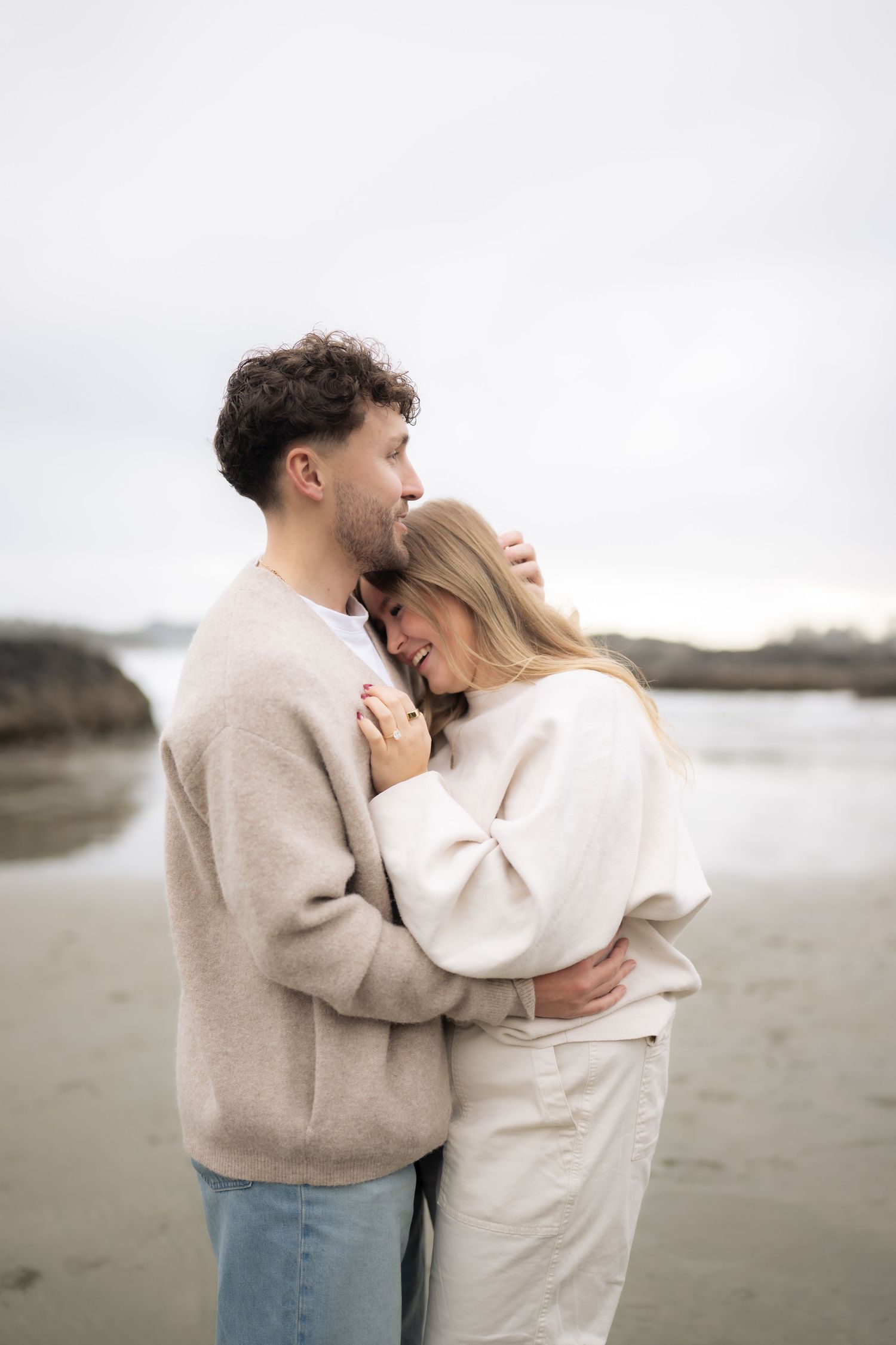 An intimate embrace between two people in neutral colored sweaters on a moody beach.