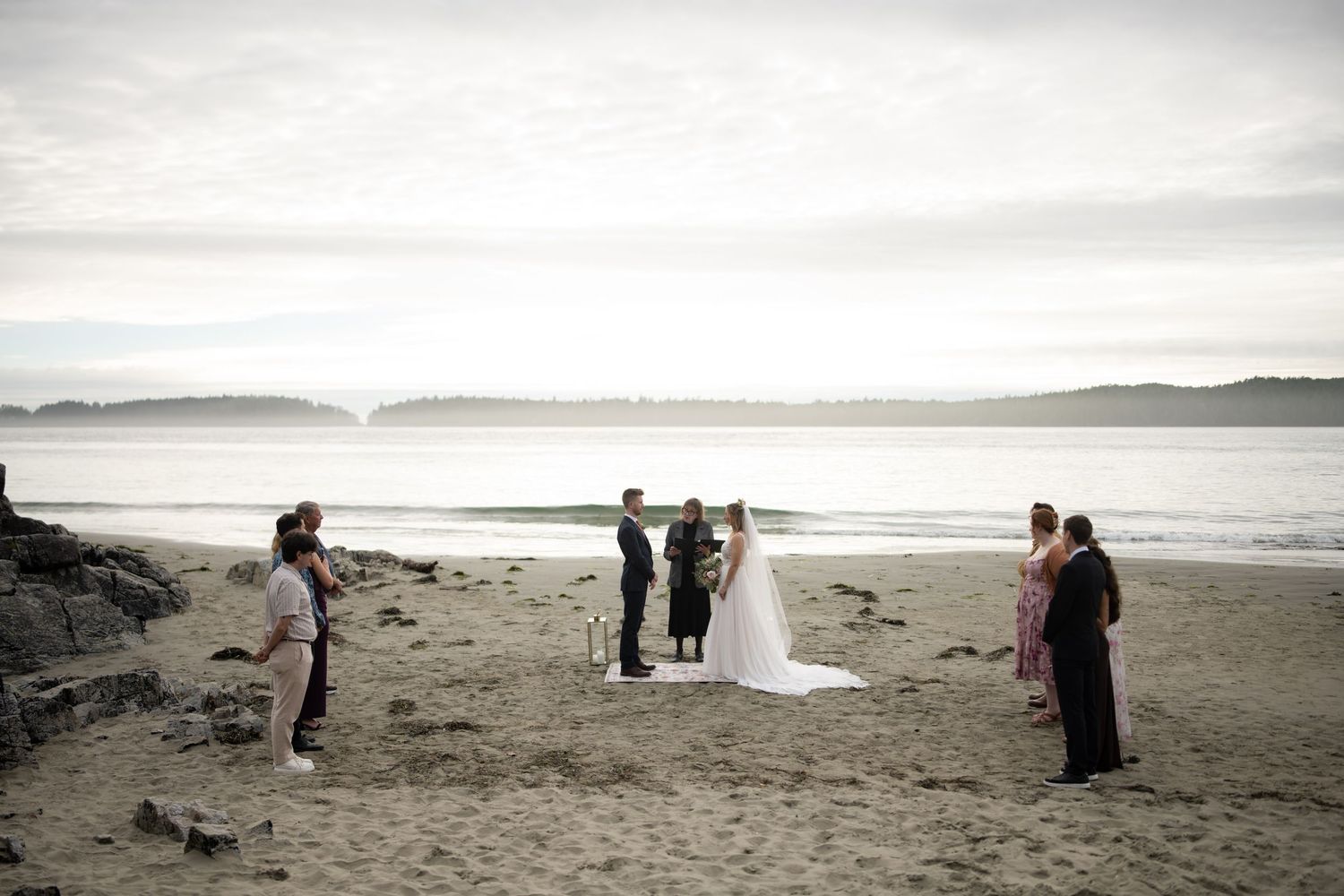 Wedding guests gather on a misty beach with mountains in the distance.