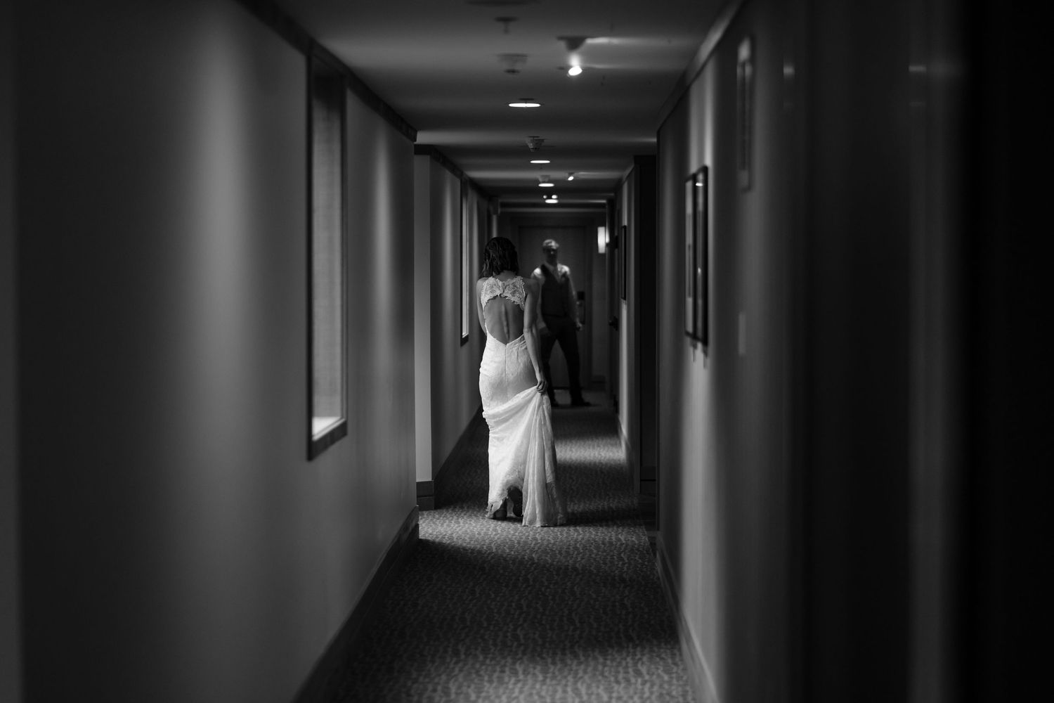 A couple walks down a dimly lit hotel hallway in formal attire.