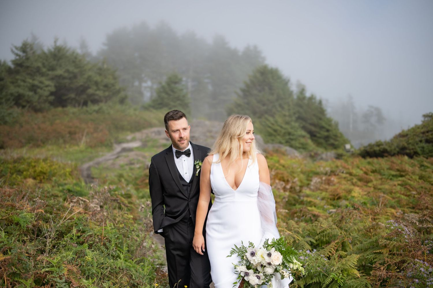 A couple in wedding attire pose on a misty mountainside trail among evergreens.