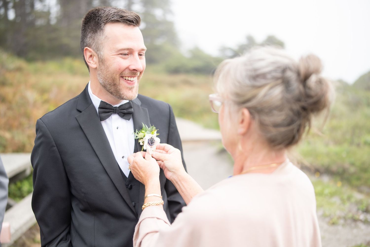 A boutonniere is being pinned onto a black tuxedo with bow tie during a wedding preparation.