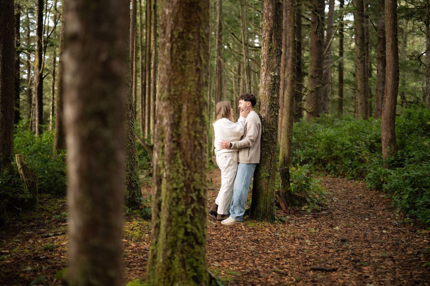 Romantic forest photoshoot with couple standing between tall evergreen trees.