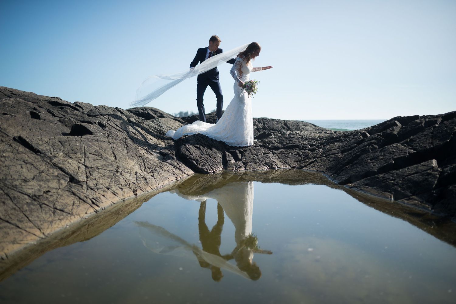 Wedding couple's reflection in still water against rocky landscape.