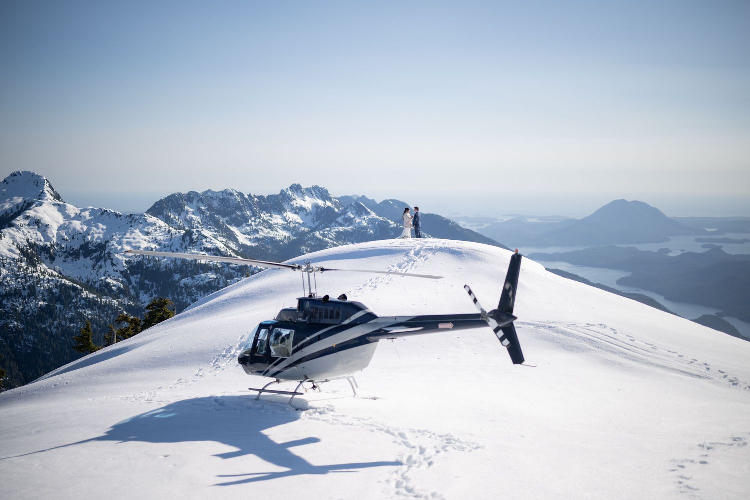 Helicopter landed on snowy mountain peak with vast mountain landscape in background.