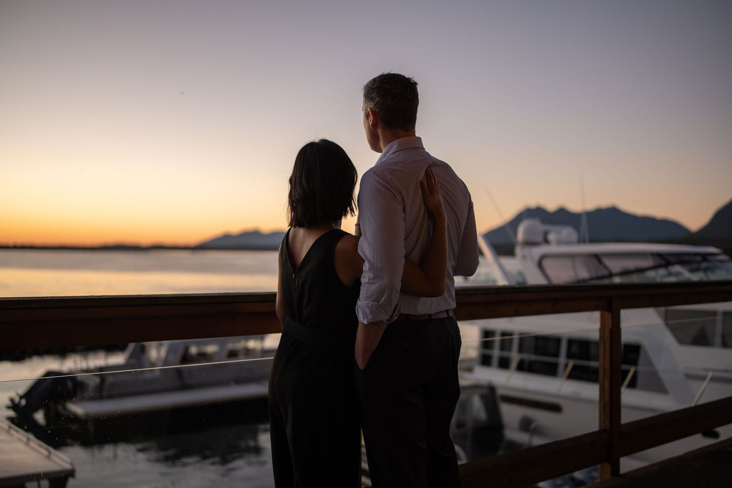 Silhouettes of guests enjoying sunset views on a waterfront deck with boats in the background.