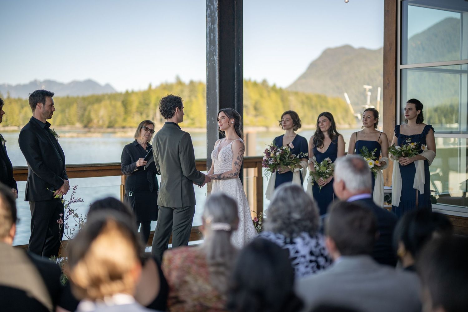 Wedding ceremony gathering with mountain and water views through large windows.