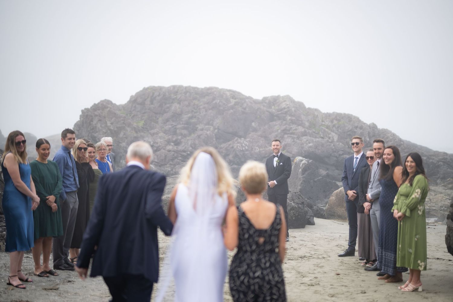 An intimate beach wedding ceremony with guests forming an aisle on rocky terrain.