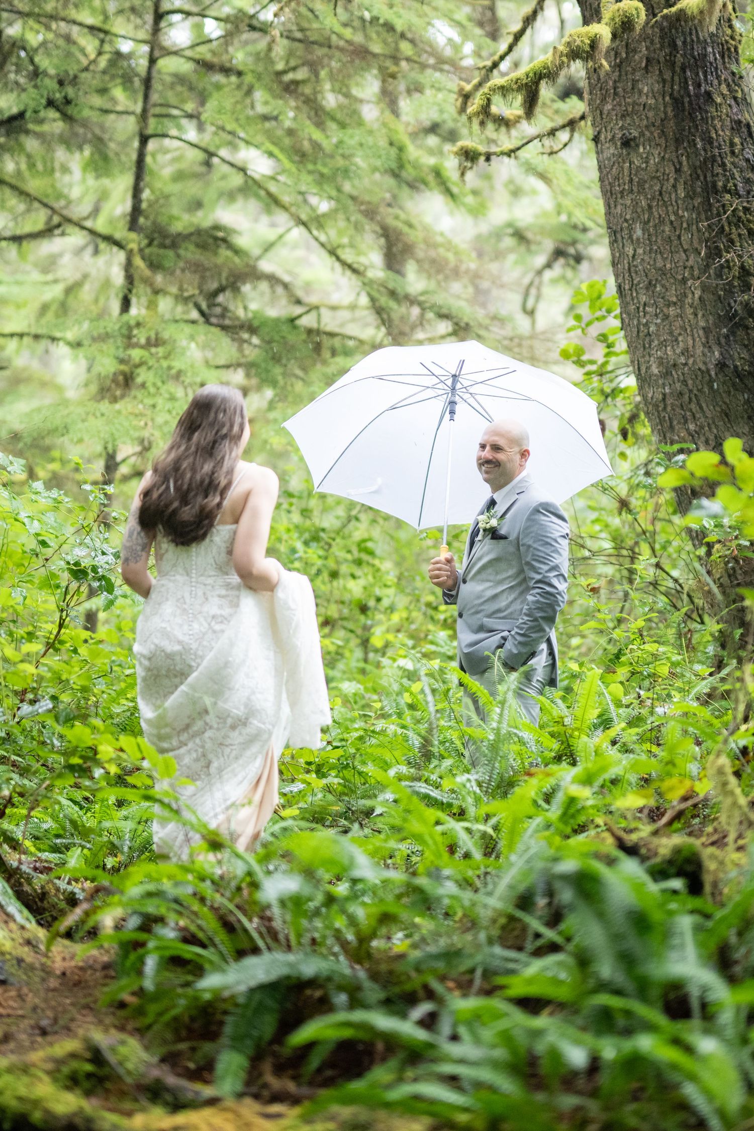 A couple shares a romantic moment under white umbrellas in a lush forest setting.