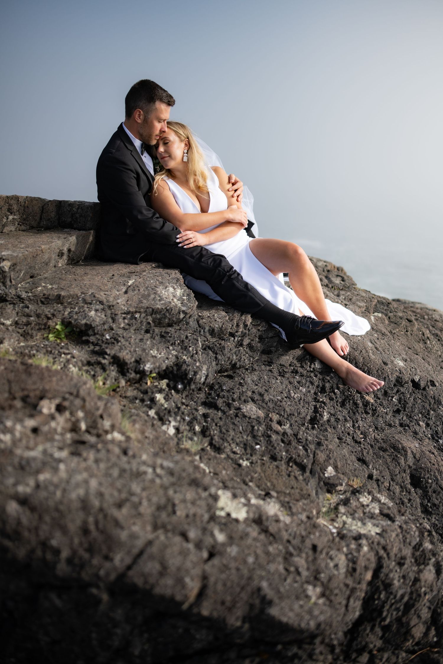 Newlyweds share romantic moments while sitting on dramatic coastal rocks.
