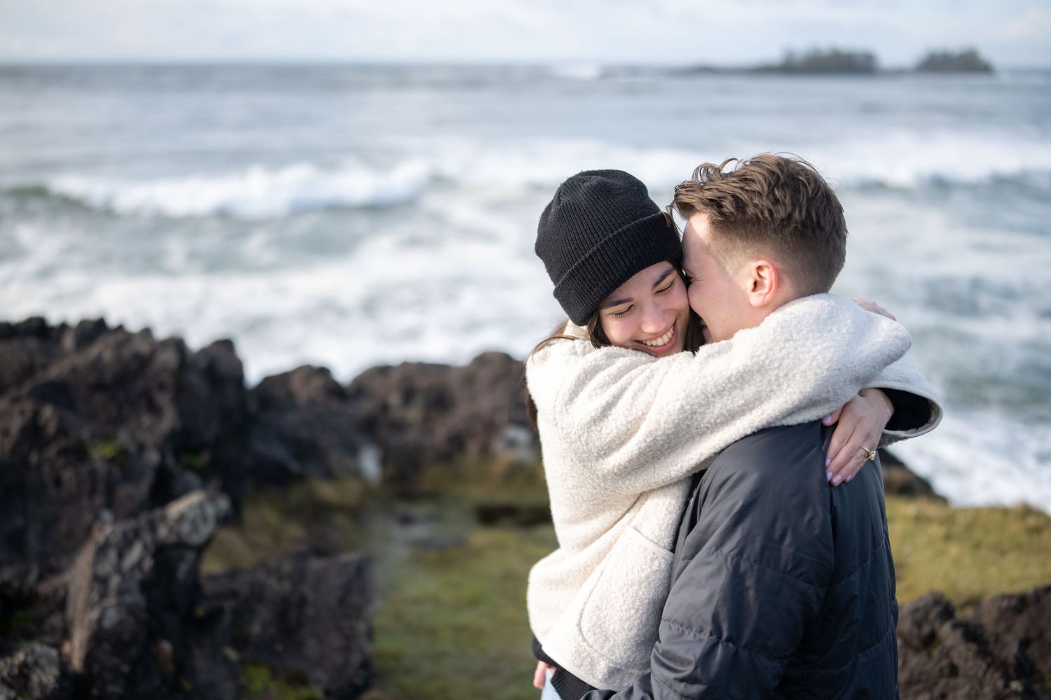 Couple embracing on a misty Pacific Northwest beach with waves and rocks in the background.