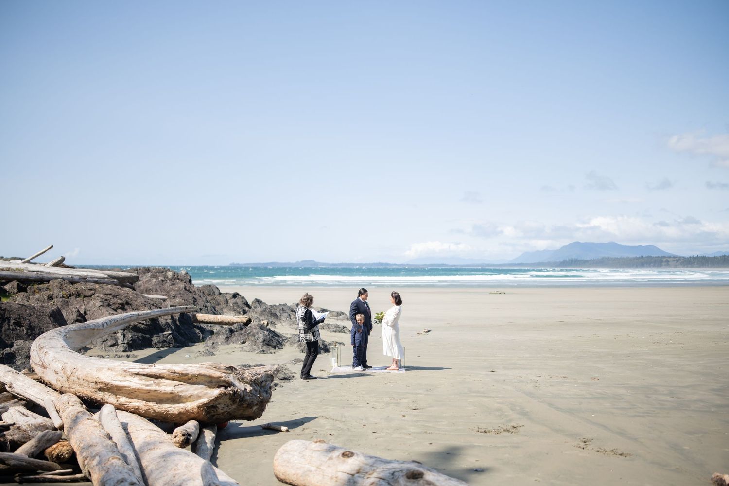 A scenic beach wedding ceremony setup with driftwood and rocks along the shoreline on a sunny day.