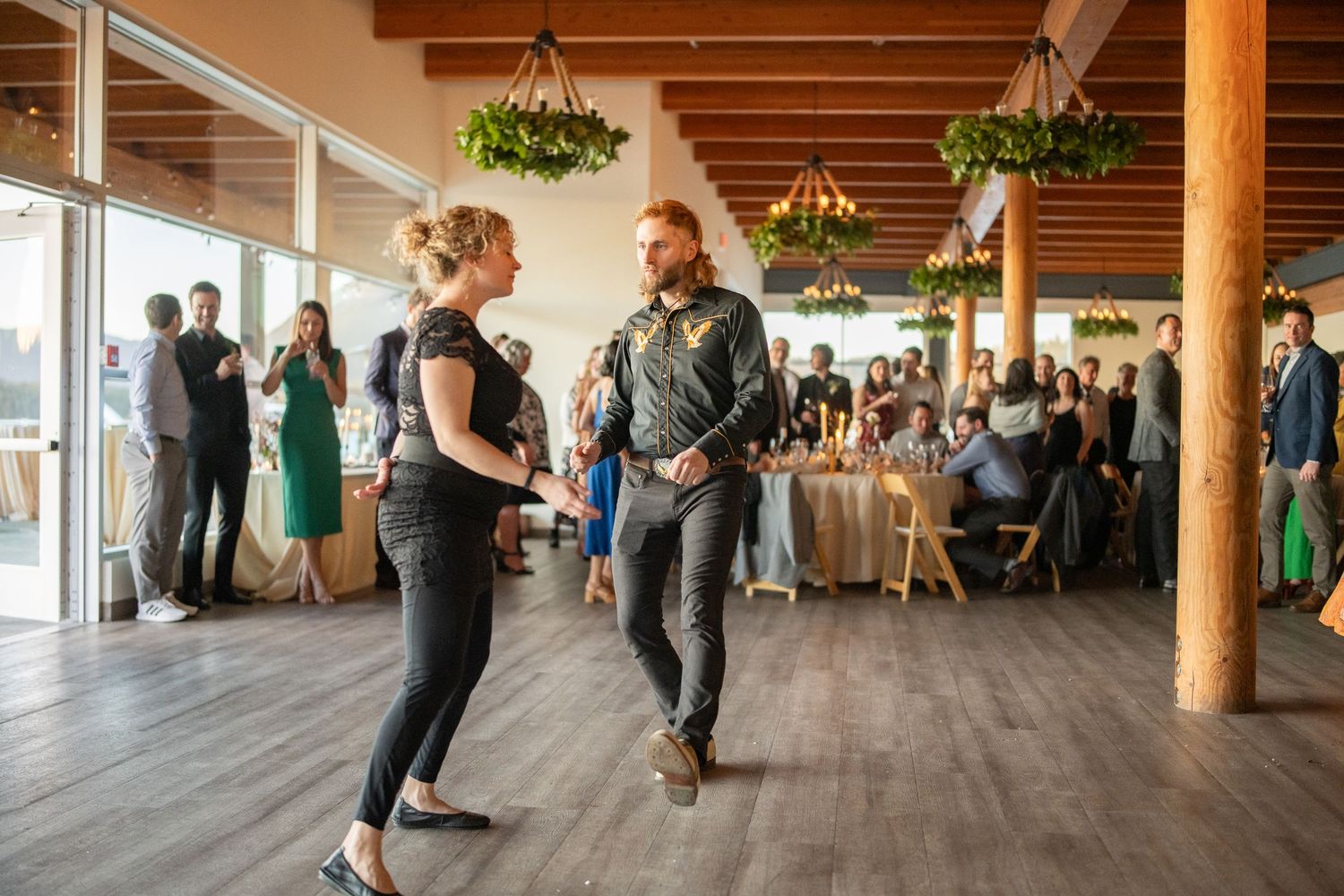 Wedding guests dancing on wooden floor under rustic beams and string lights.