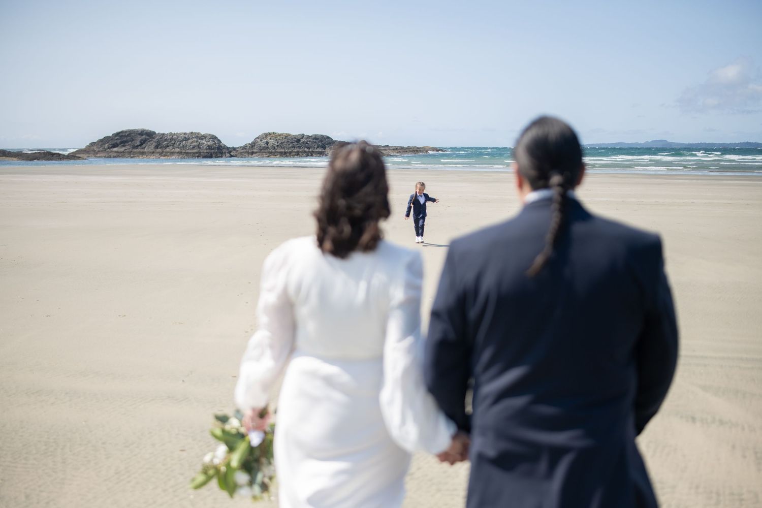 Wedding couple walks along scenic coastal beach with mountains in the distance.