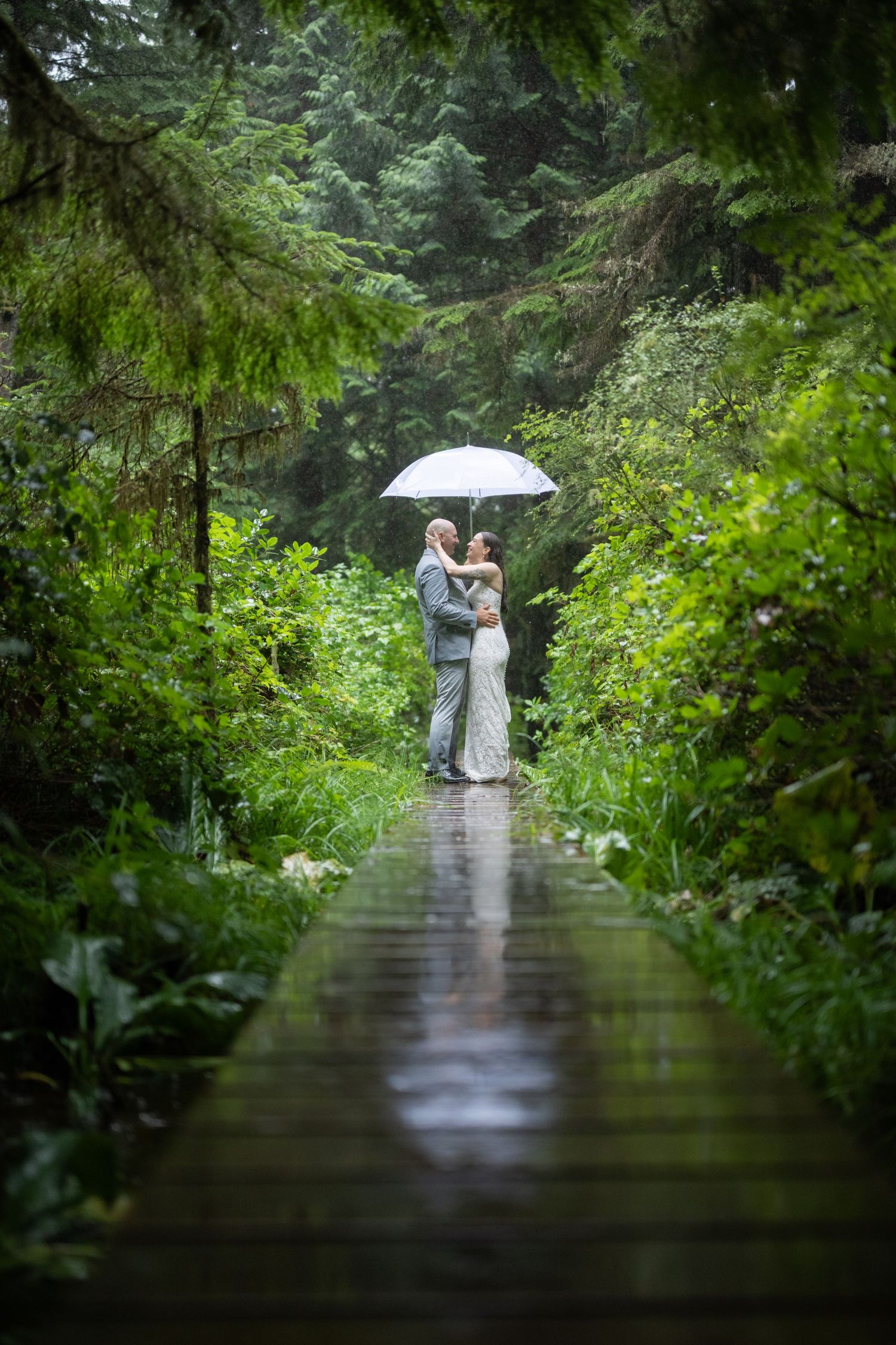 A couple embraces on a wooden boardwalk in a rainy forest while holding a white umbrella.