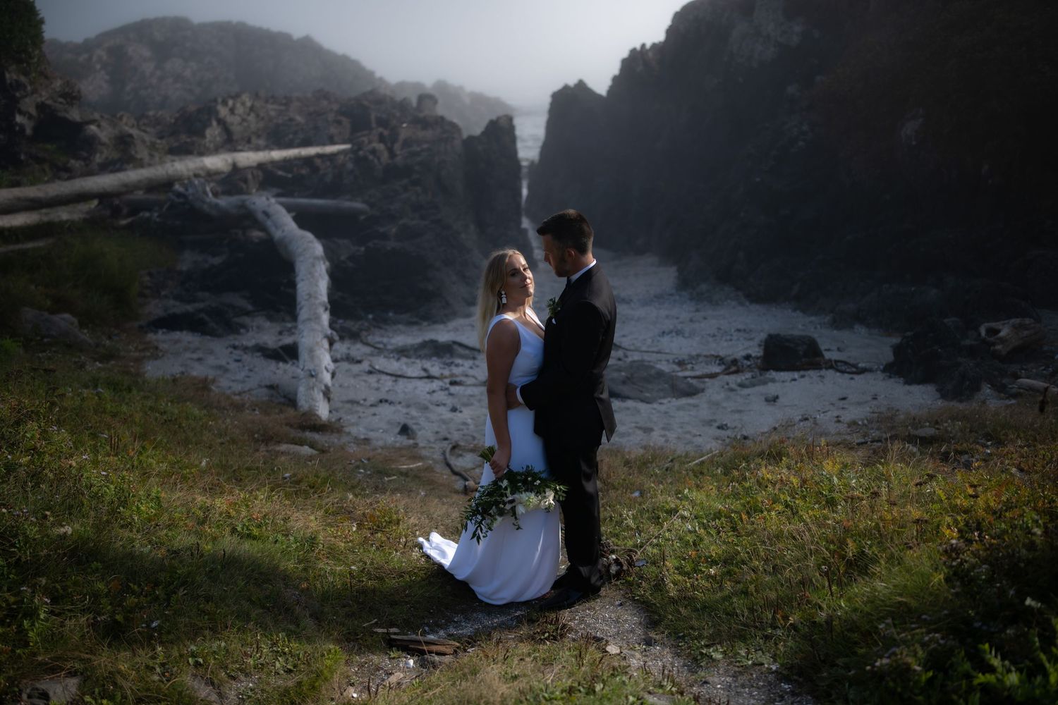 Couple poses on dramatic rocky coastline with misty cliffs in background.