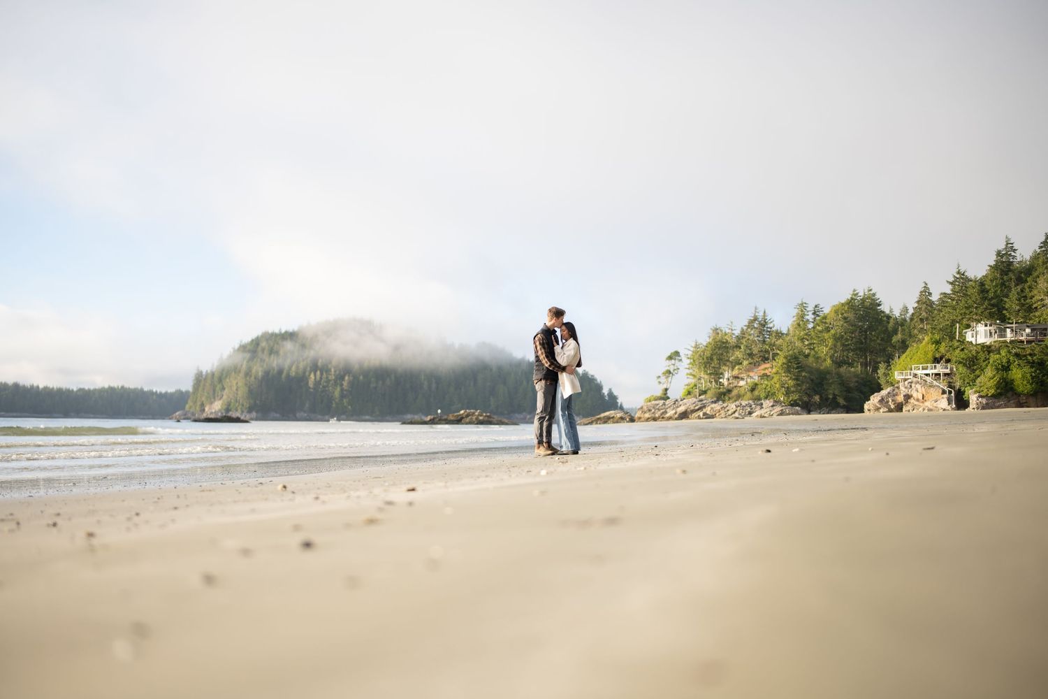 Couple embracing on empty beach with misty mountains in background.