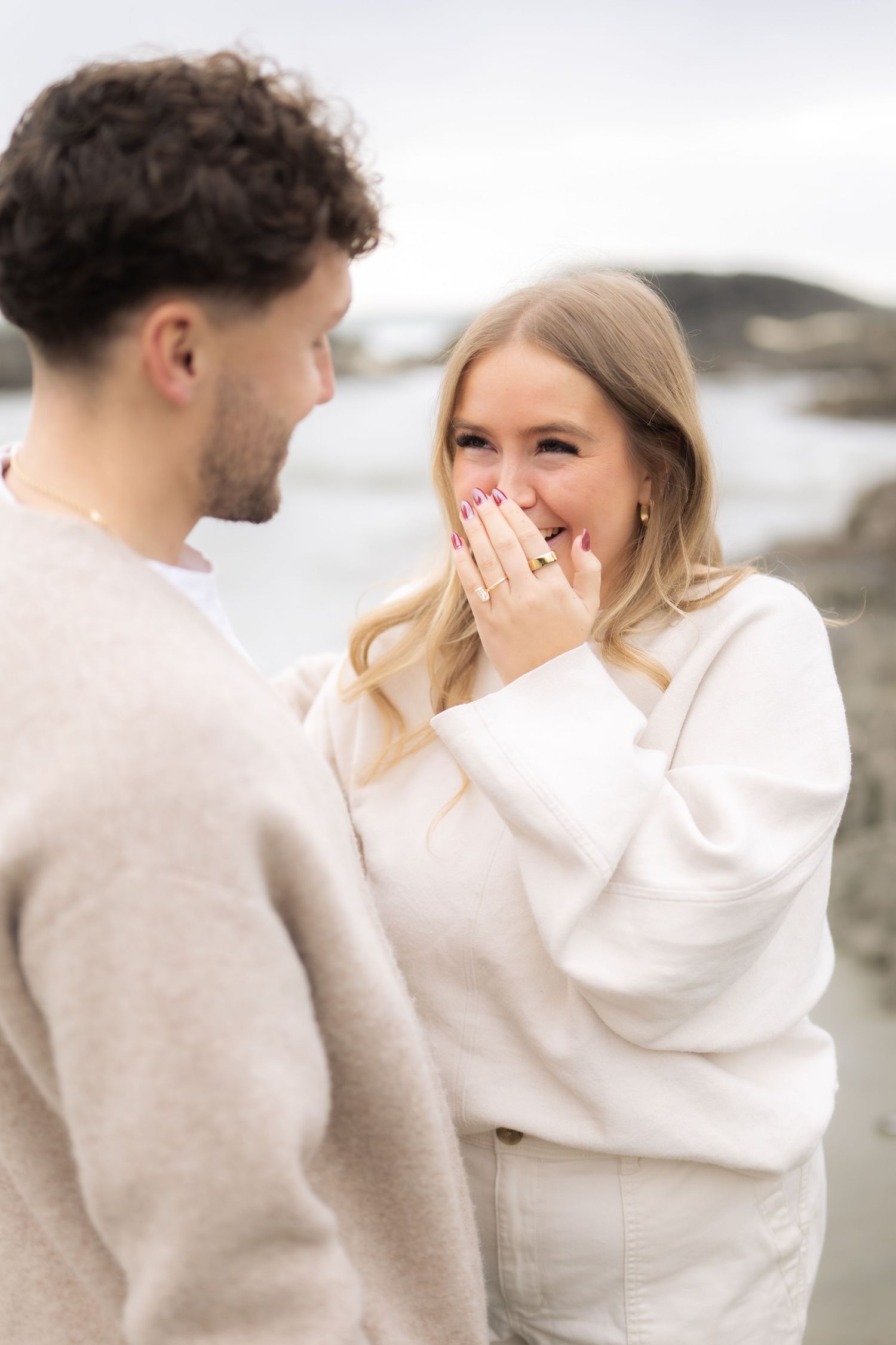 Sweet candid reaction during a surprise beach proposal with rocky coastline backdrop.