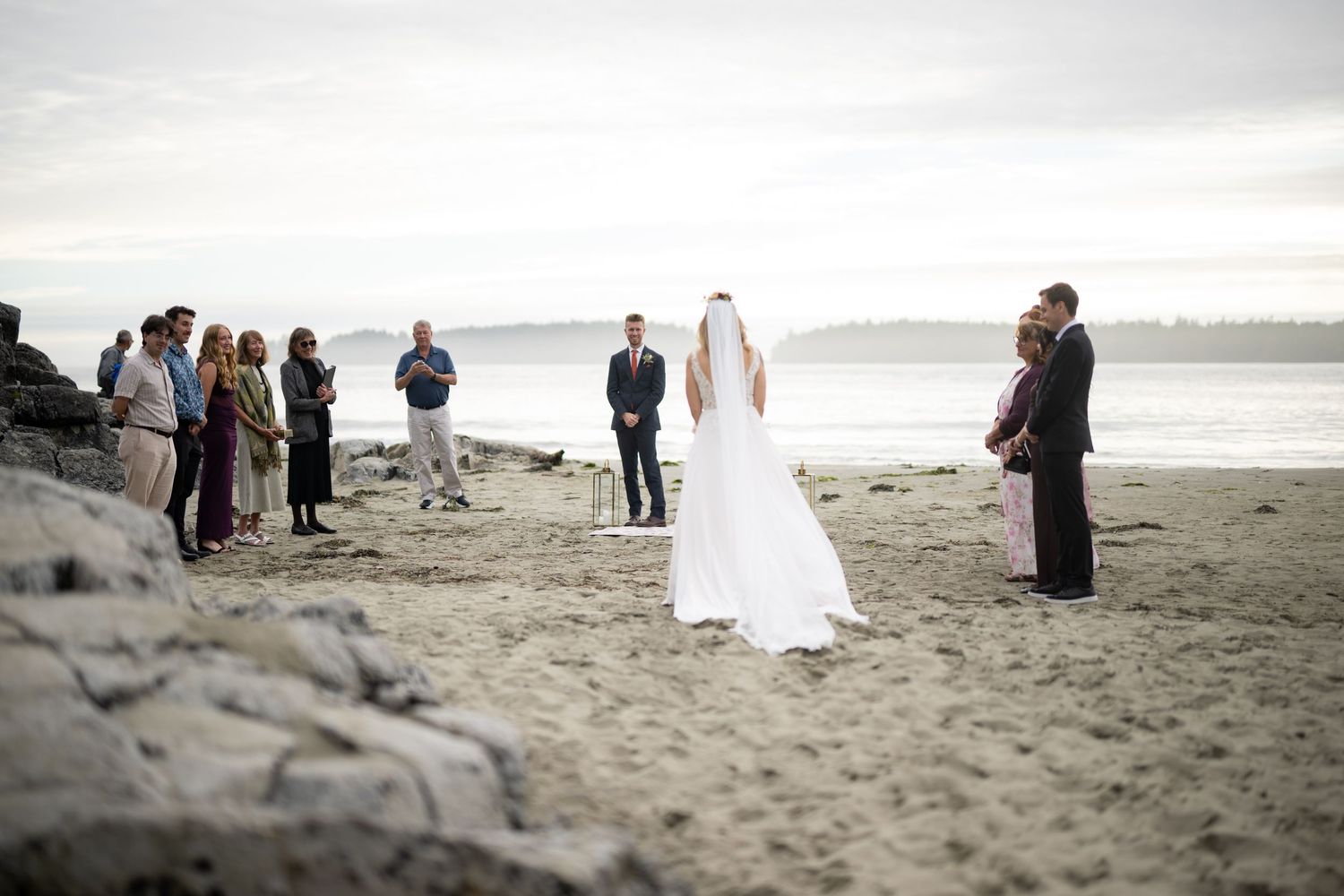 Wedding ceremony attendees gather on a cloudy beach with distant islands visible on the horizon.