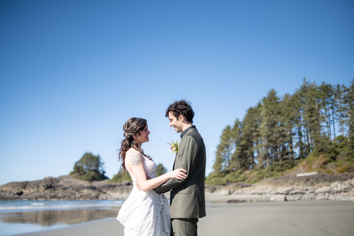 A couple shares an intimate moment on a scenic beach with evergreen trees in the background.