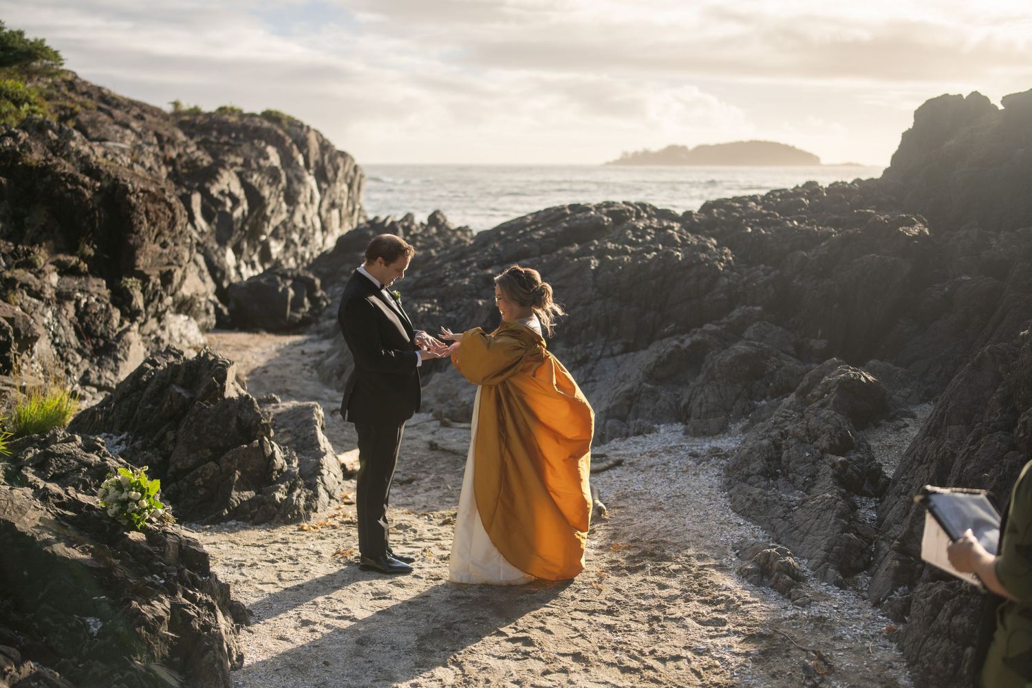 Two people sit on rocky coastal cliffs overlooking the ocean at sunset.