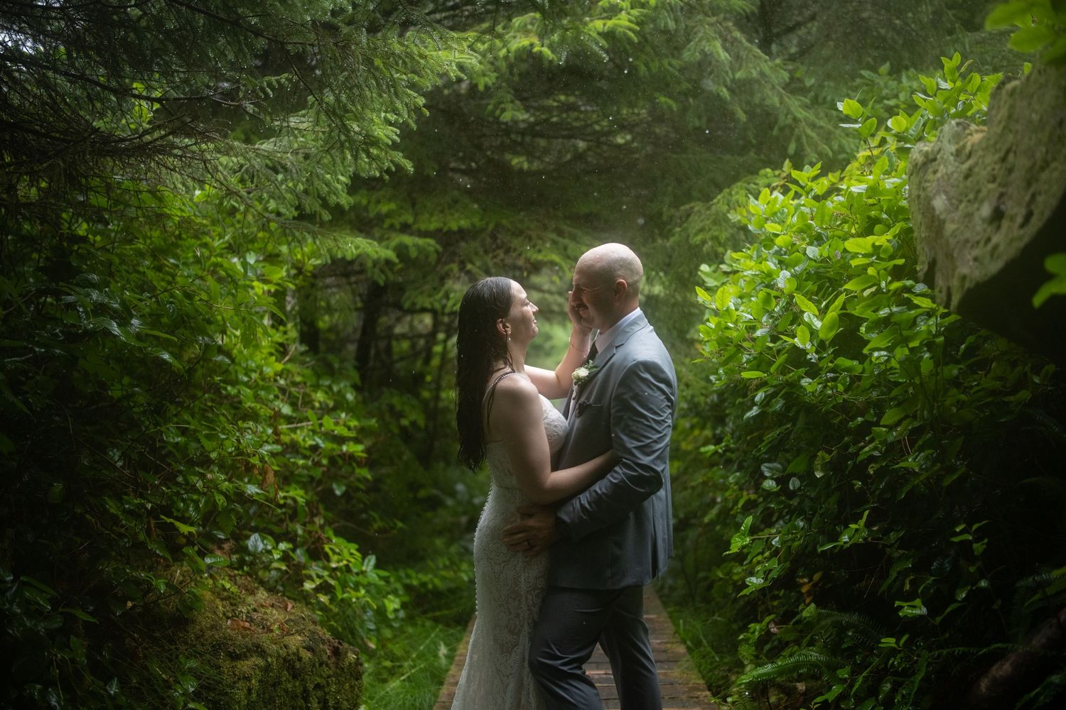 Newlyweds share a romantic moment on a forest path surrounded by green foliage.