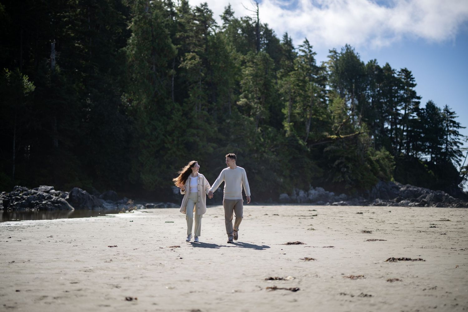 Two people walking hand in hand along a scenic beach with mountains and forest in the background.