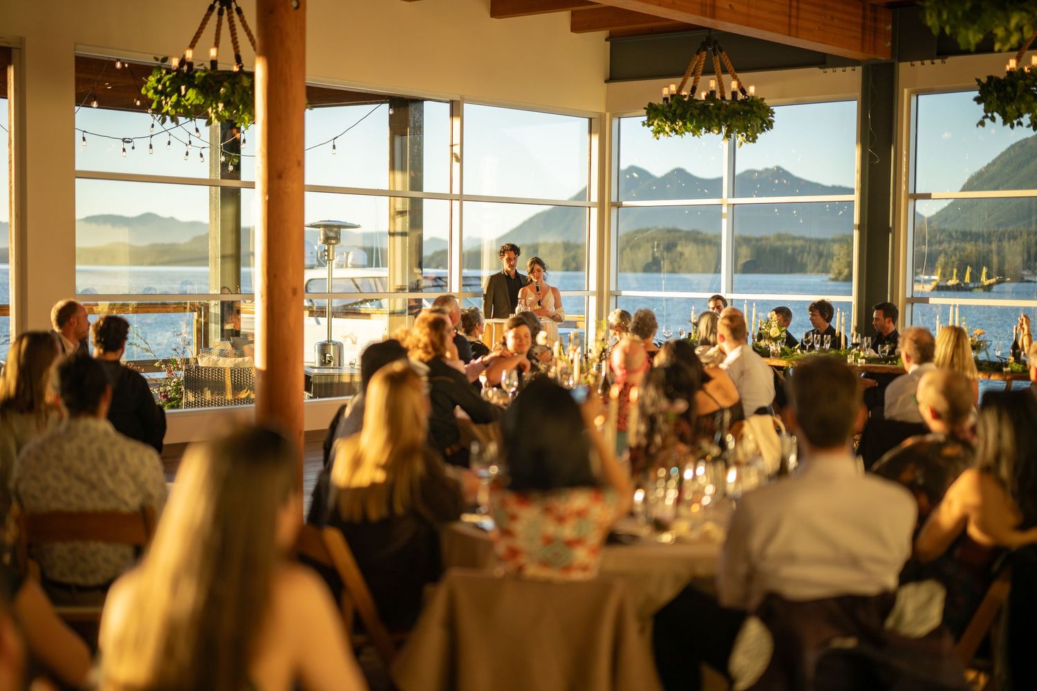 Spacious waterfront venue with exposed beams and floor-to-ceiling windows overlooking mountains.