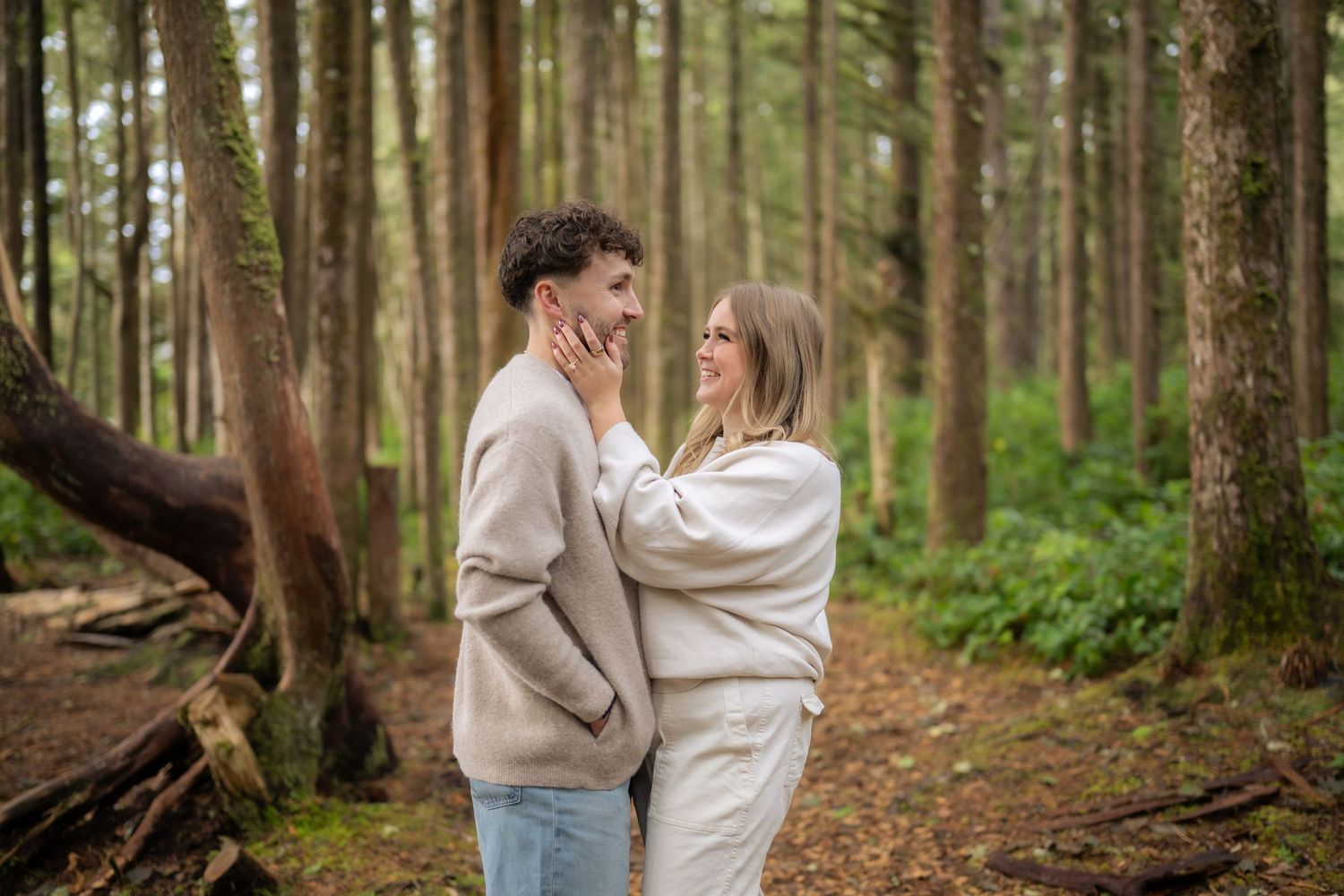 Couple embracing among tall pine trees on forest floor covered in needles.