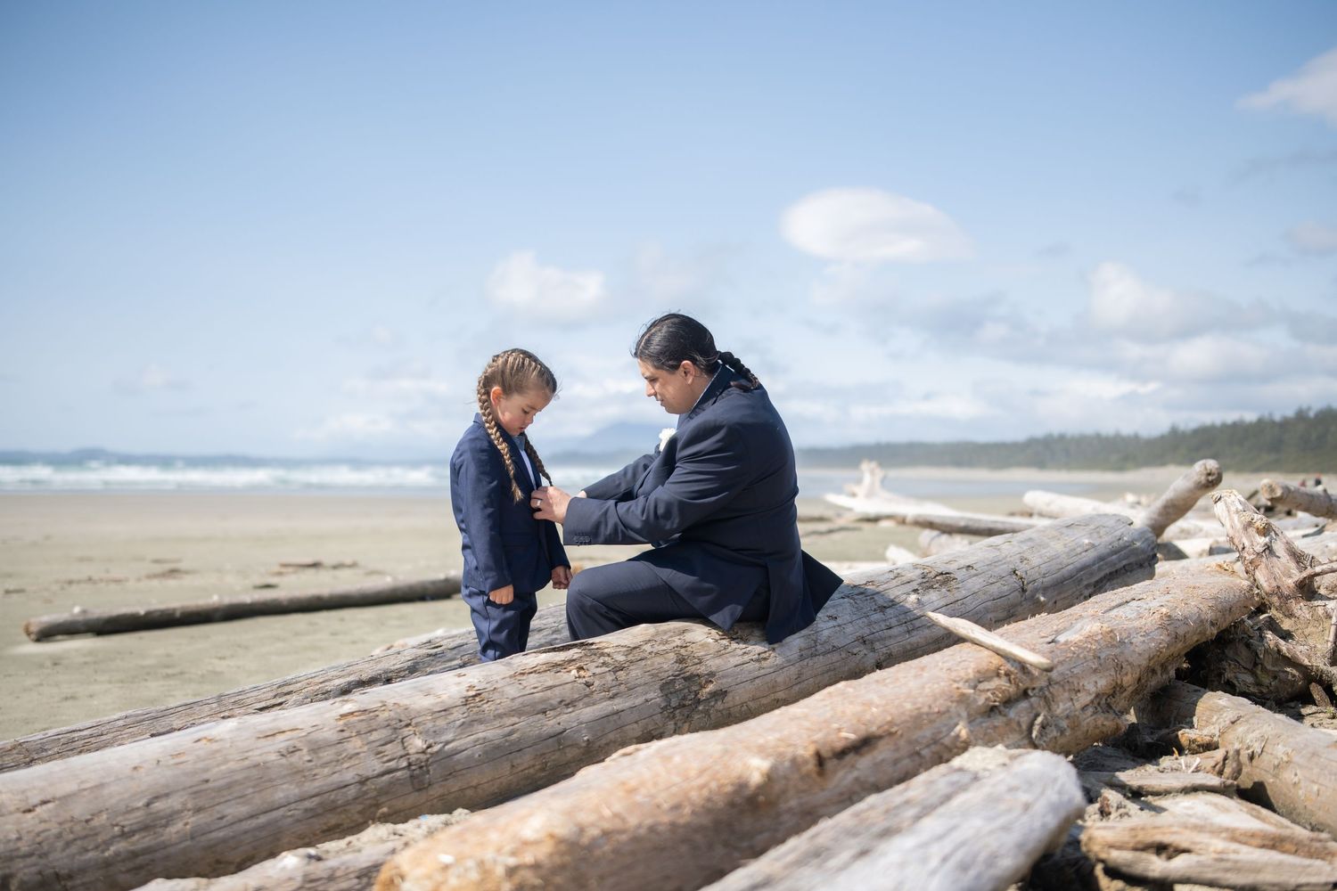 A couple sits together on a large driftwood log at a scenic beach with mountains in the background.
