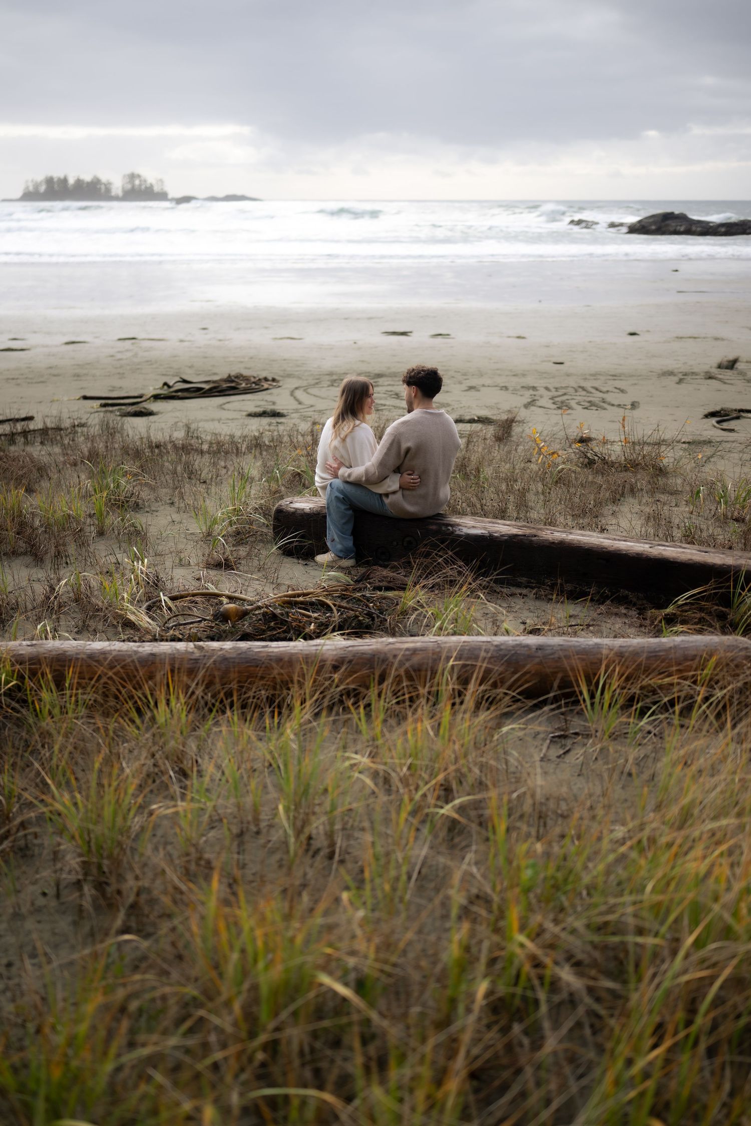 Couple sitting on driftwood log at misty beach with waves and coastal grasses in foreground.