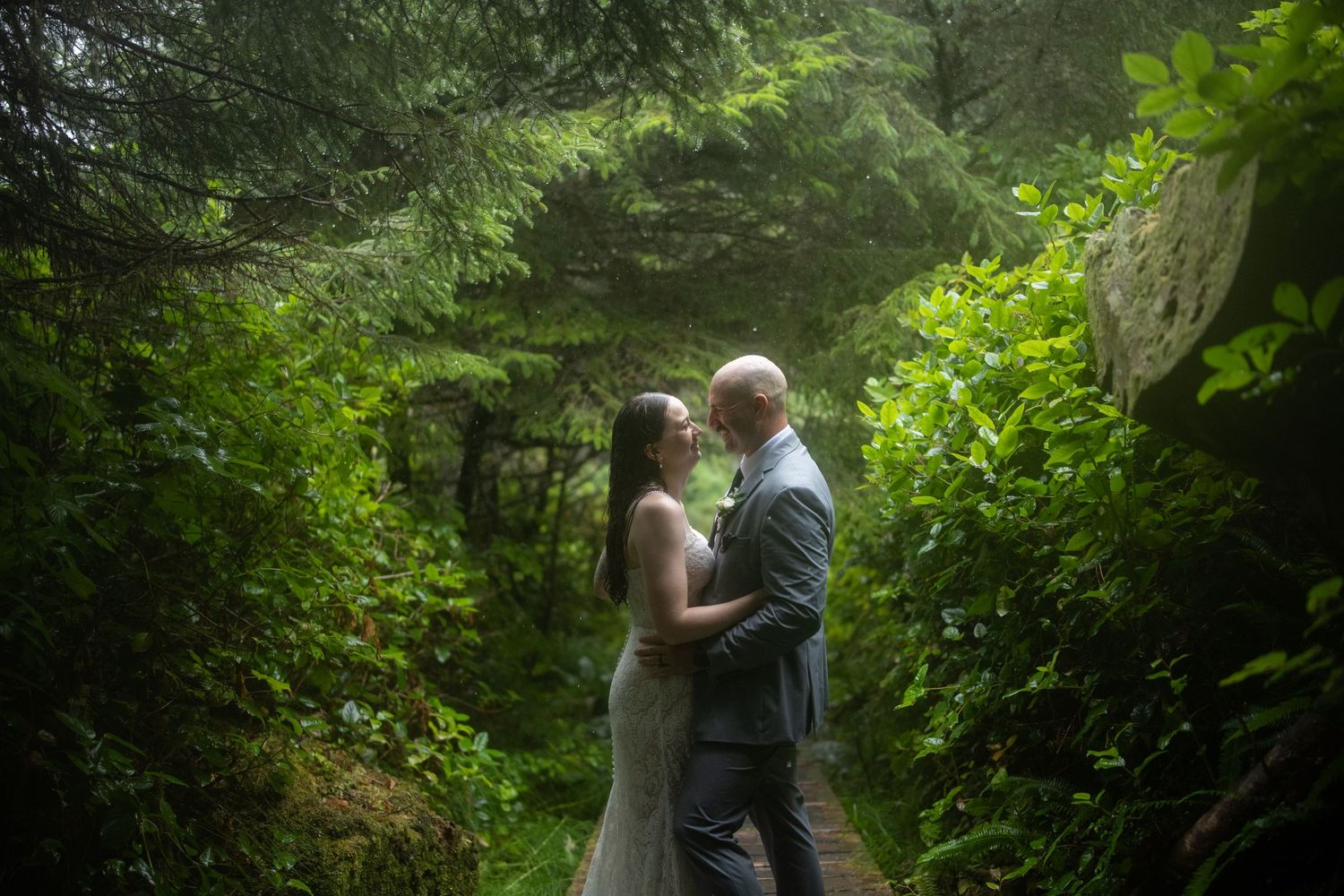 Wedding couple embraces on a wooded pathway surrounded by evergreen trees.