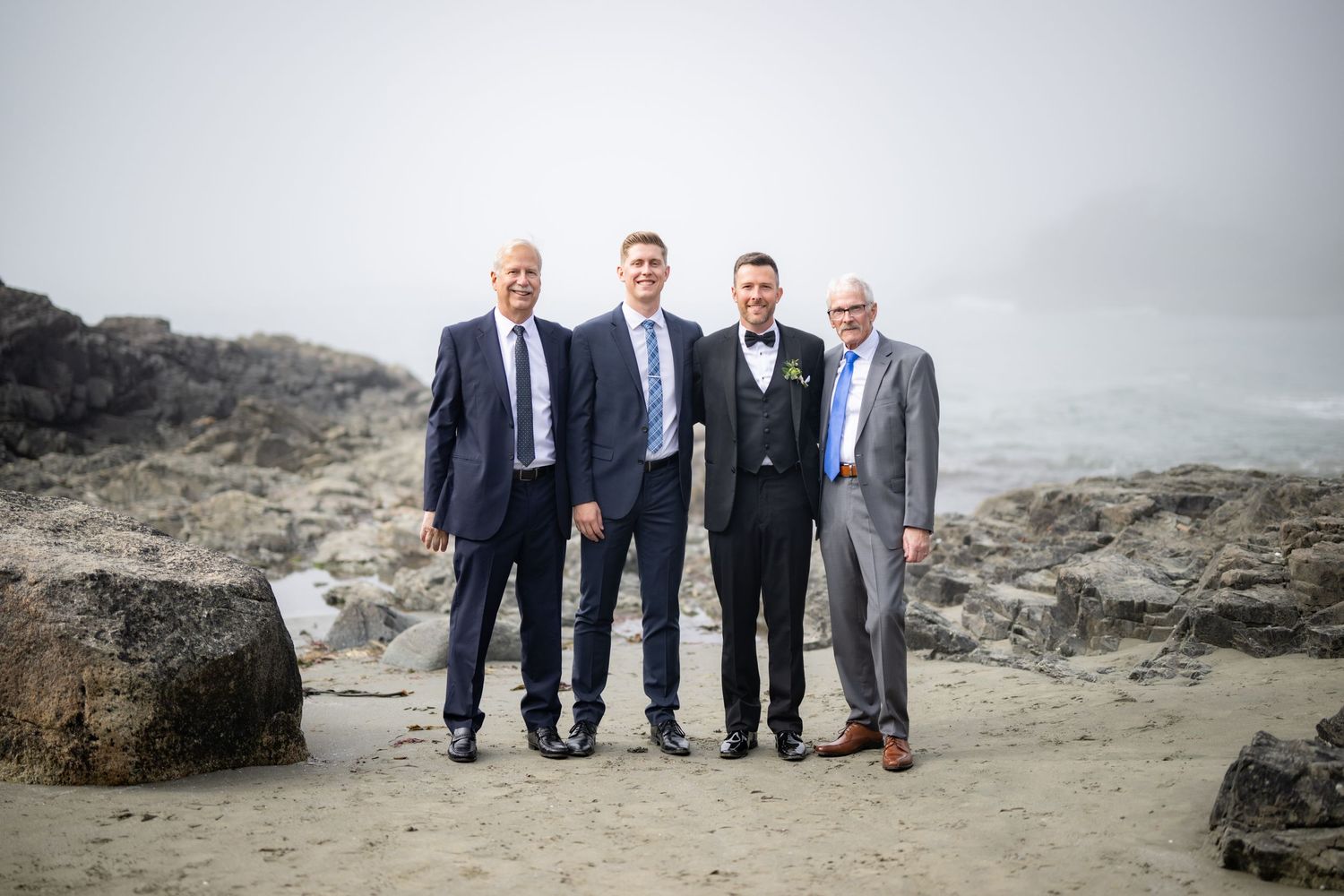 Groomsmen in dark suits pose together on rocky coastal landscape.
