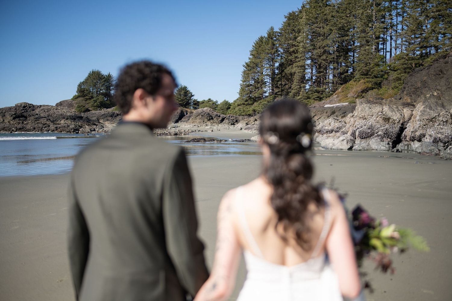 A couple stands together on a scenic beach with pine trees and rocky cliffs.