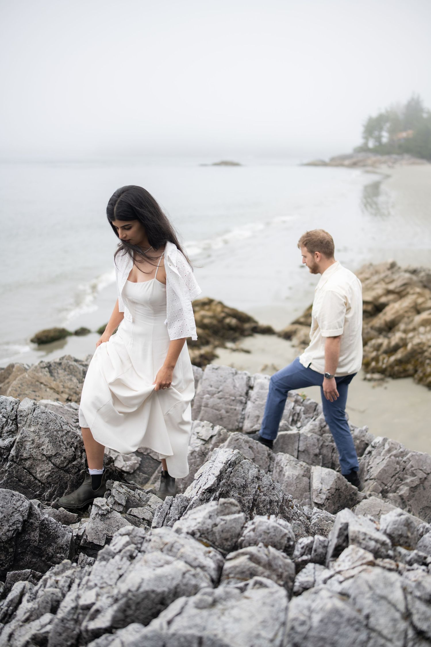 Two people navigate rocky coastal terrain while wearing white and denim outfits.