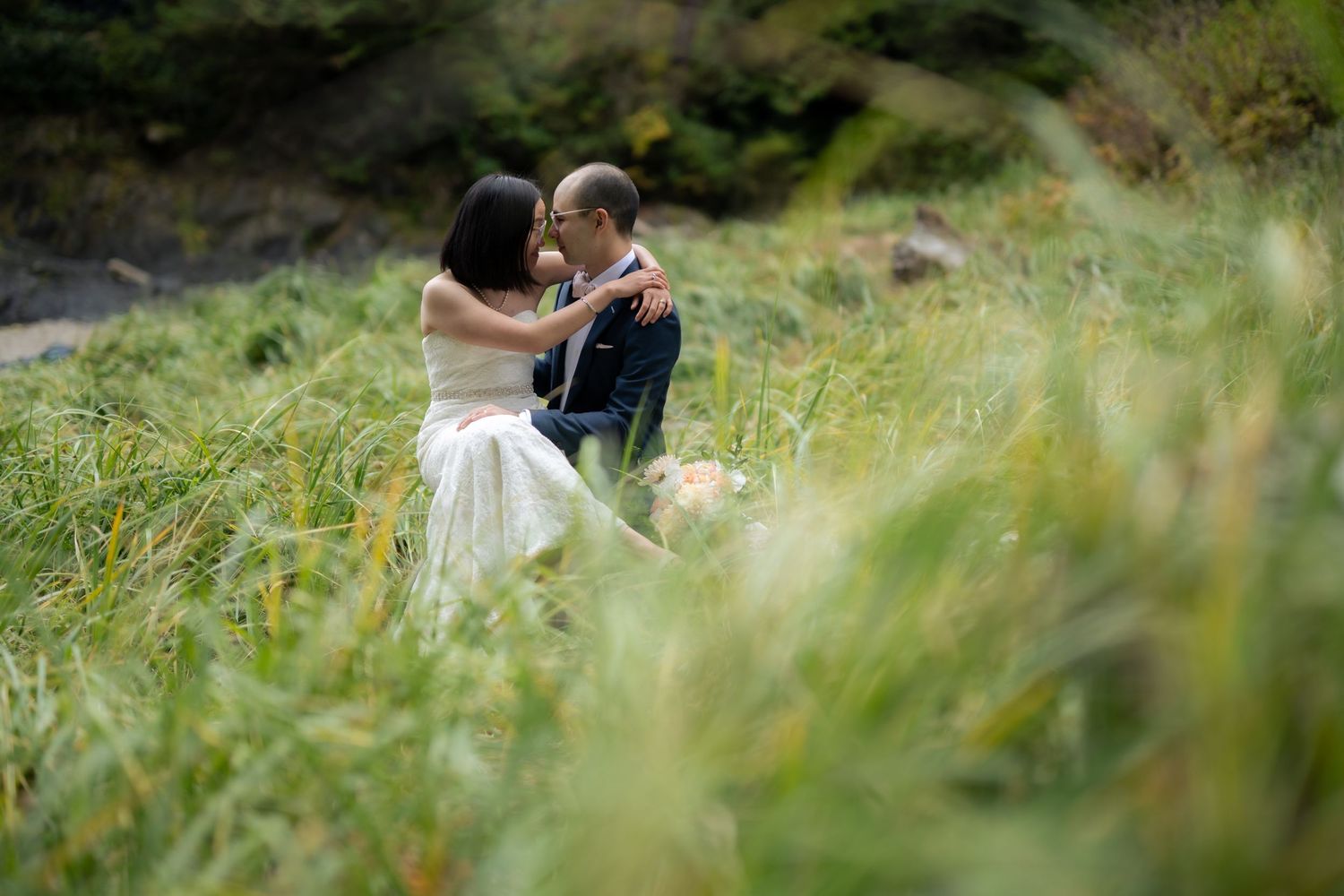Wedding couple photographed through swaying coastal grasses near ocean cliffs.