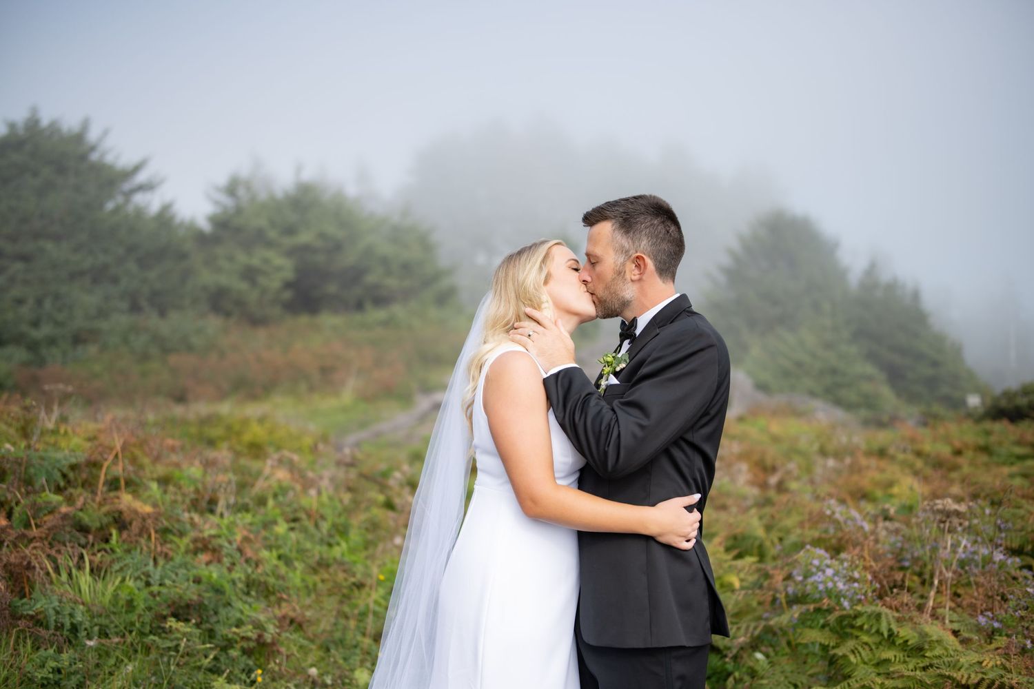 A romantic moment captured on a foggy mountainside with greenery in the background.