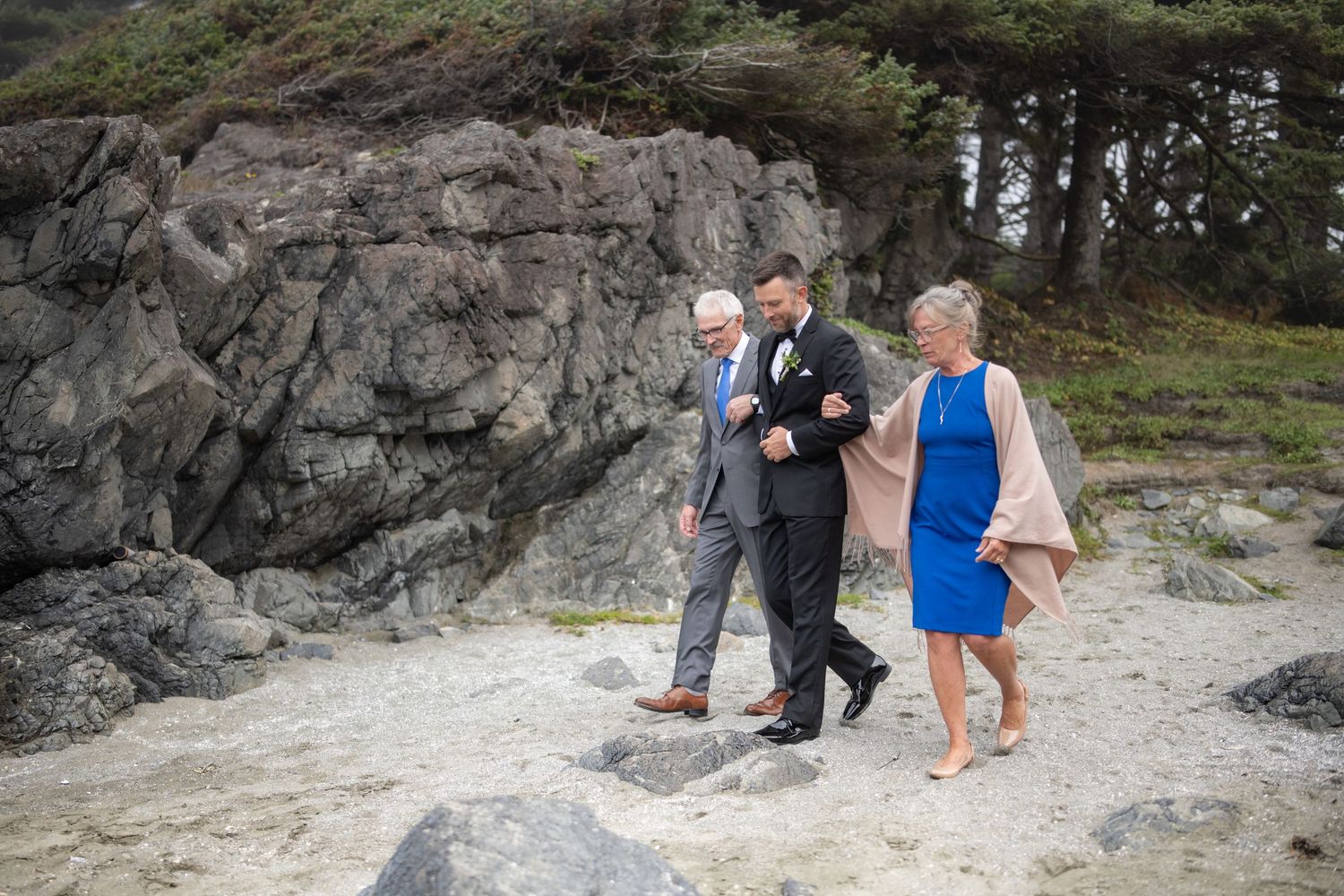 Wedding party members walk along a rocky coastal path on a foggy day.
