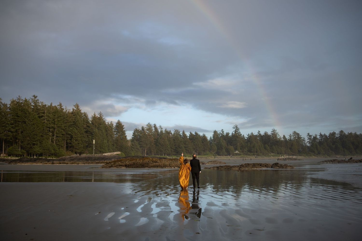 Figures stand in reflection on wet sand with evergreen forest and rainbow in background.