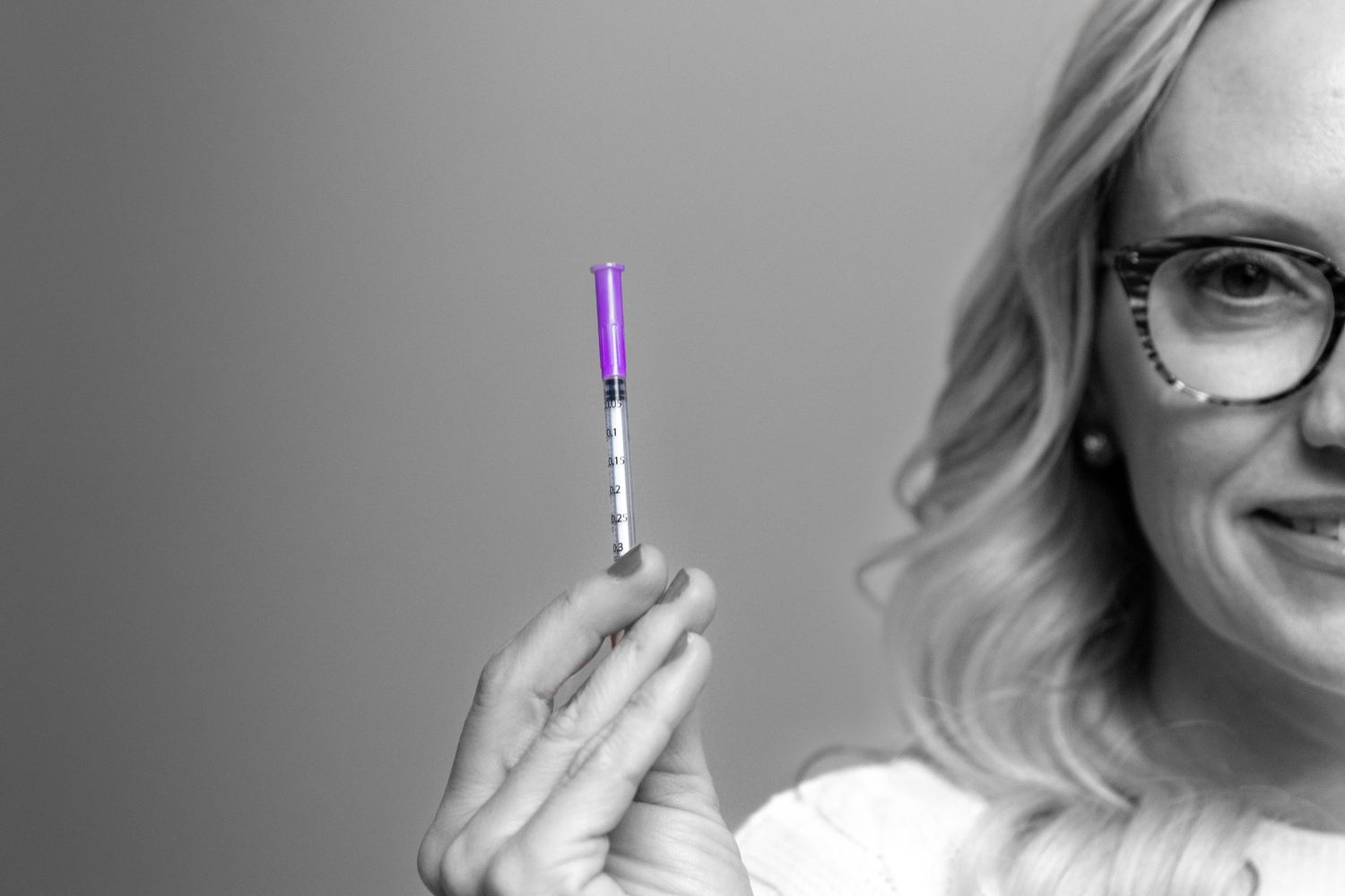Smiling woman with wavy hair and patterned glasses holds up a small insulin syringe with a purple cap in a black and white portrait.