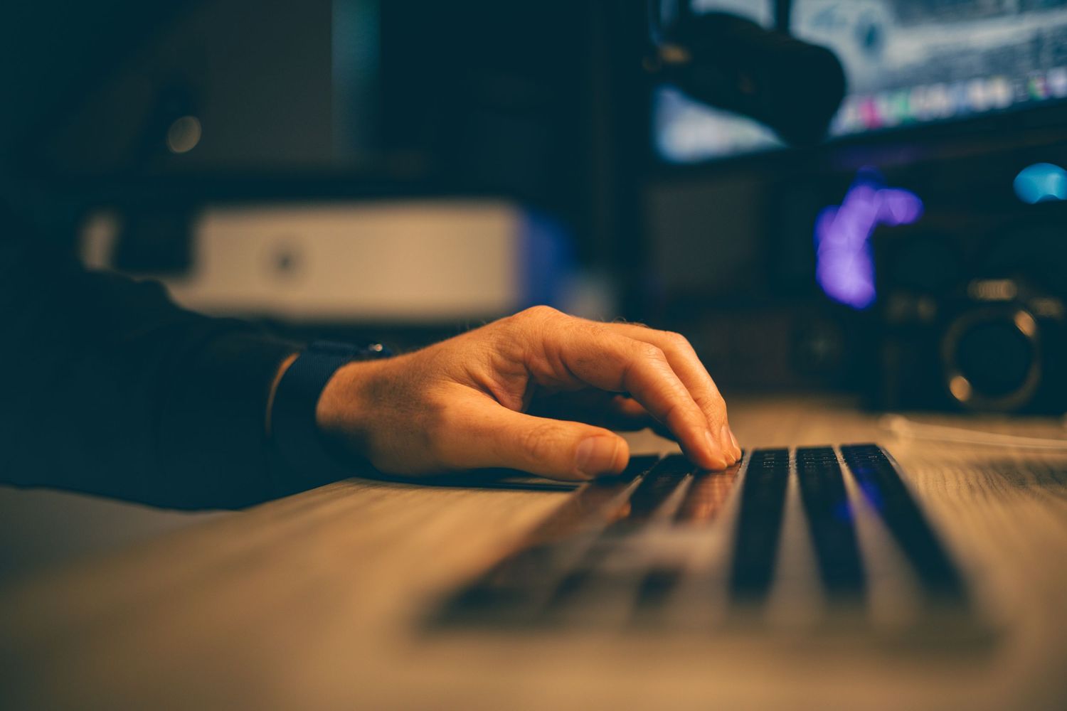 A close-up of a hand resting on a keyboard in a dimly lit video editing studio with glowing monitors in the background.