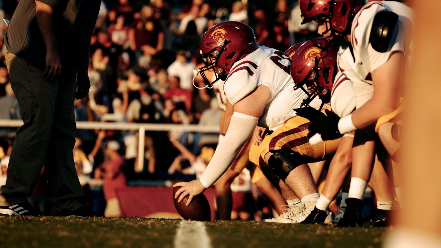 Football players in maroon and white uniforms crouch at the line of scrimmage as the center grips the ball before the snap.