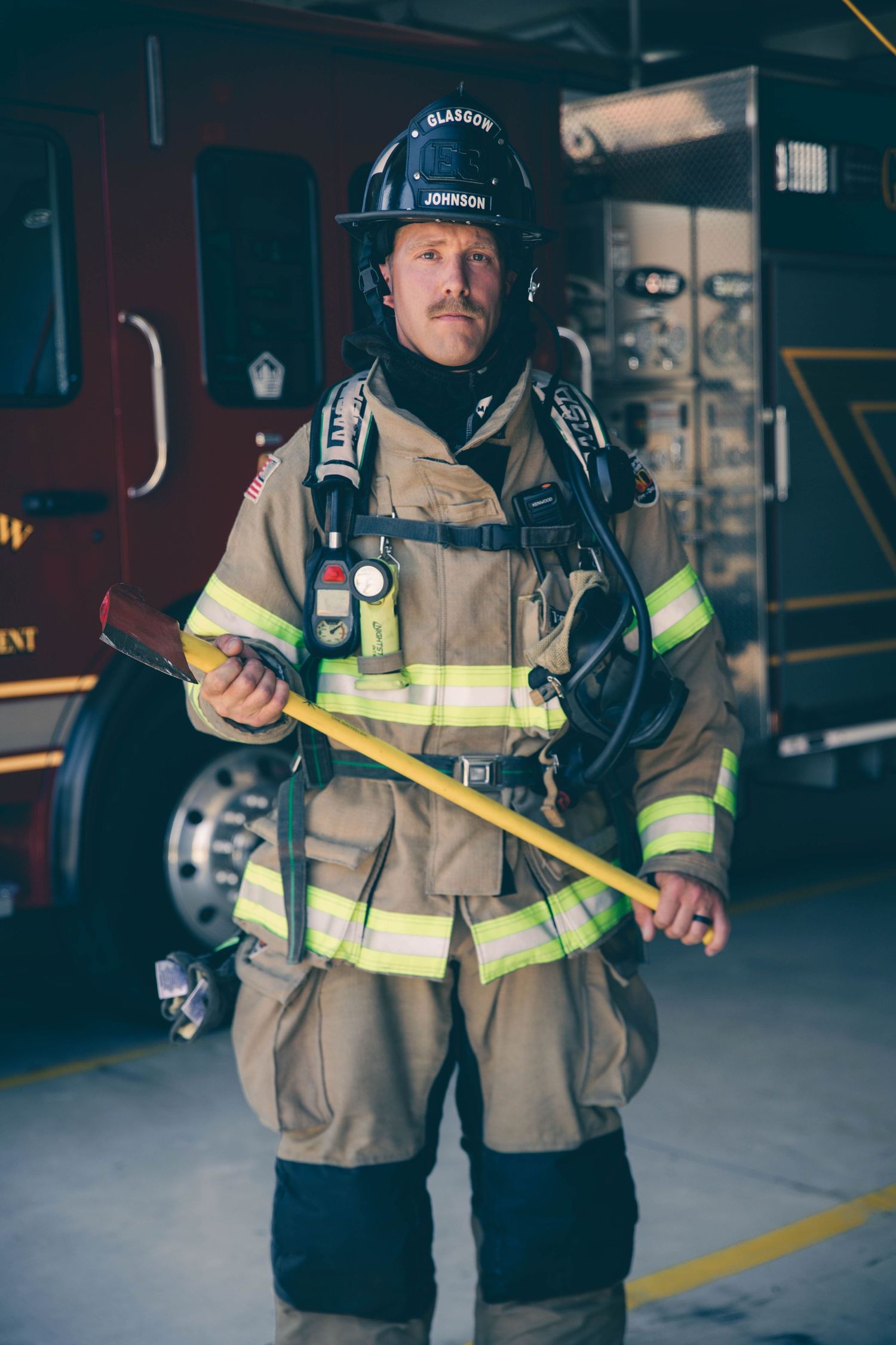 Firefighter in tan turnout gear and a black helmet holds a fire axe while standing in front of a red fire truck in the station.