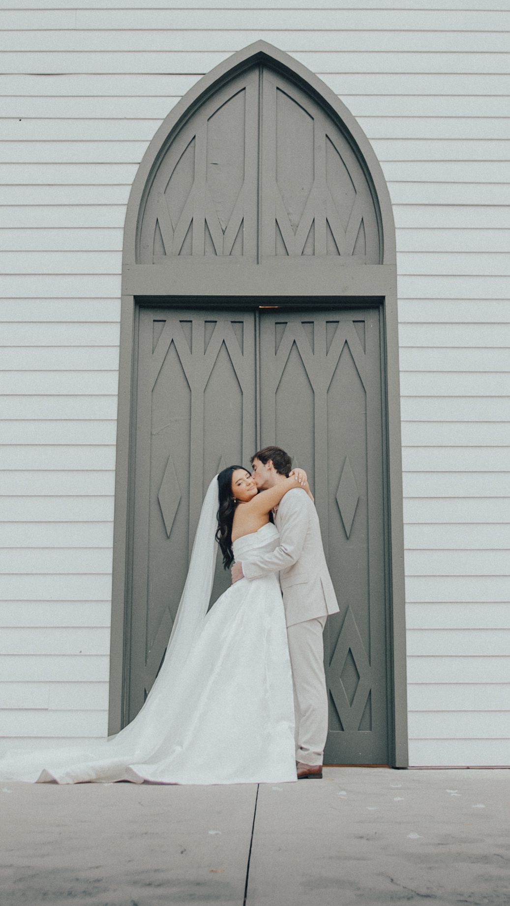 Bride in a strapless white gown and long veil kisses her groom in a light suit in front of a tall arched gray wooden door.