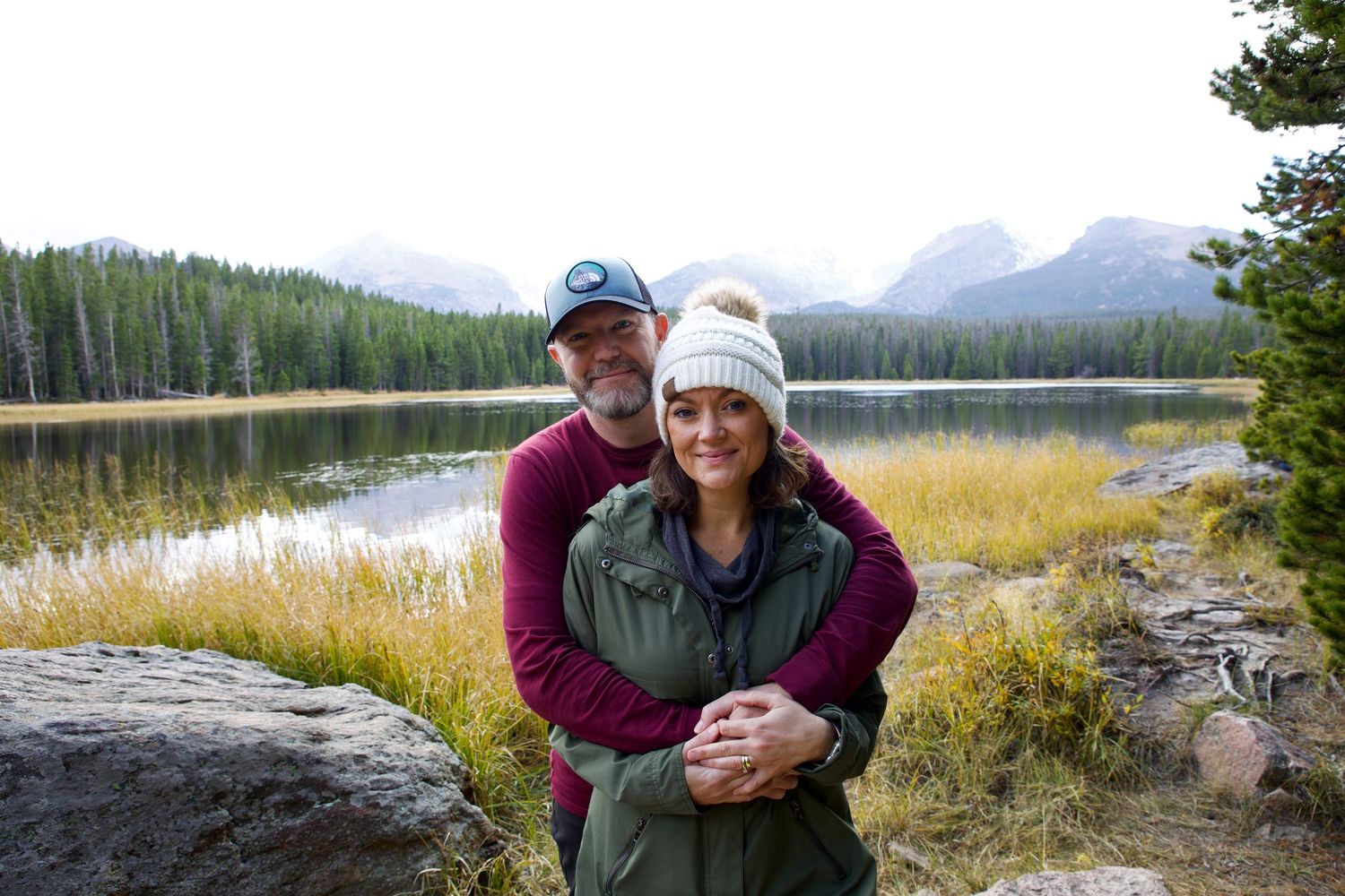 A smiling couple in beanies and jackets embrace beside a calm mountain lake with grassy shores and peaks behind them.