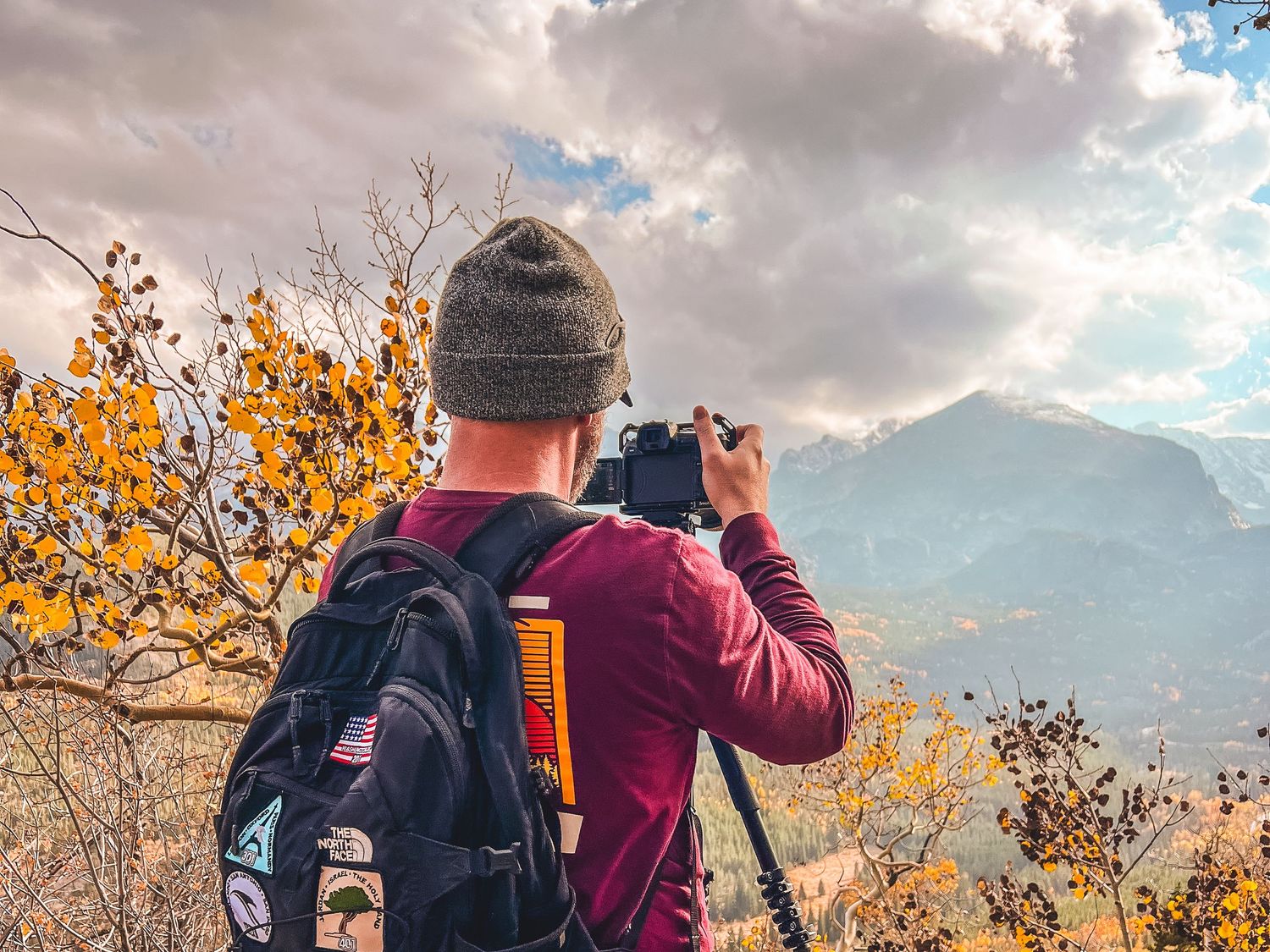 A photographer in a beanie and backpack with patches shoots a mountain view beside golden aspen trees on an autumn day.