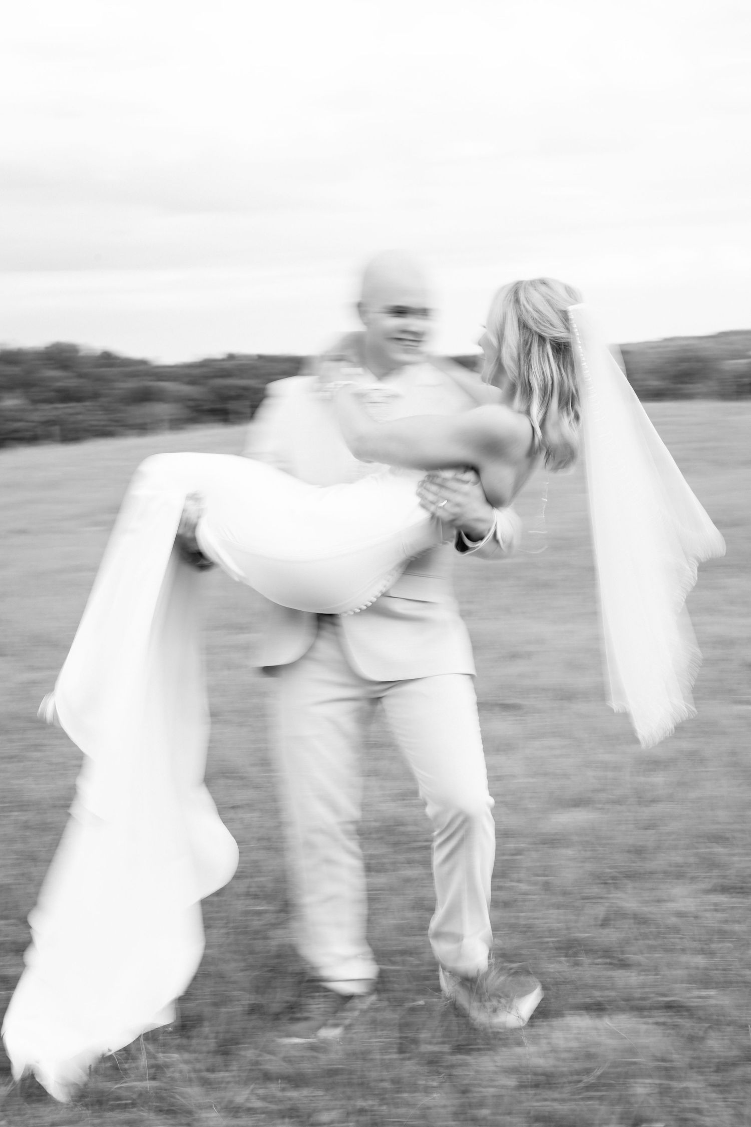 Black and white blurred motion photo of a groom in a light suit carrying his bride in a long veil across a grassy field.