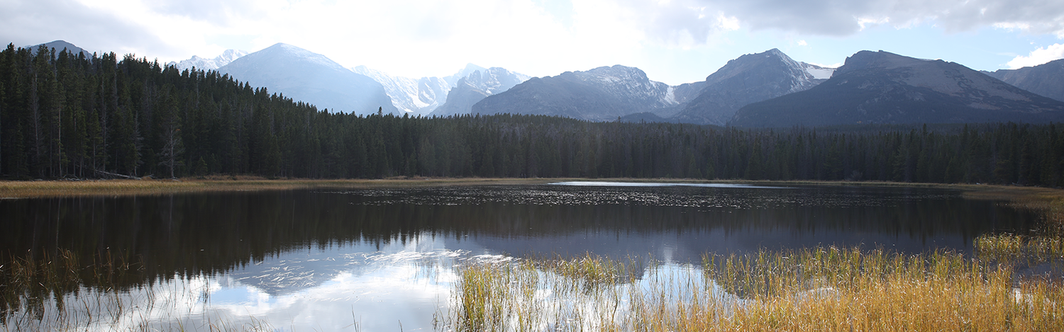 A panoramic view of rugged mountain peaks reflected in a still alpine lake bordered by dense evergreen forest.