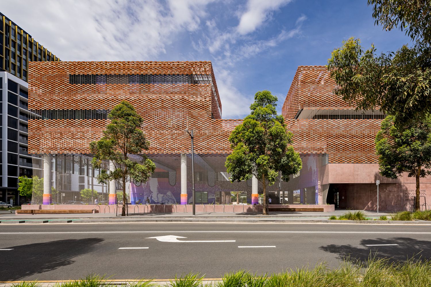 Modern terracotta brick building with a glass-fronted ground floor, pastel columns and young trees beside a quiet urban street.