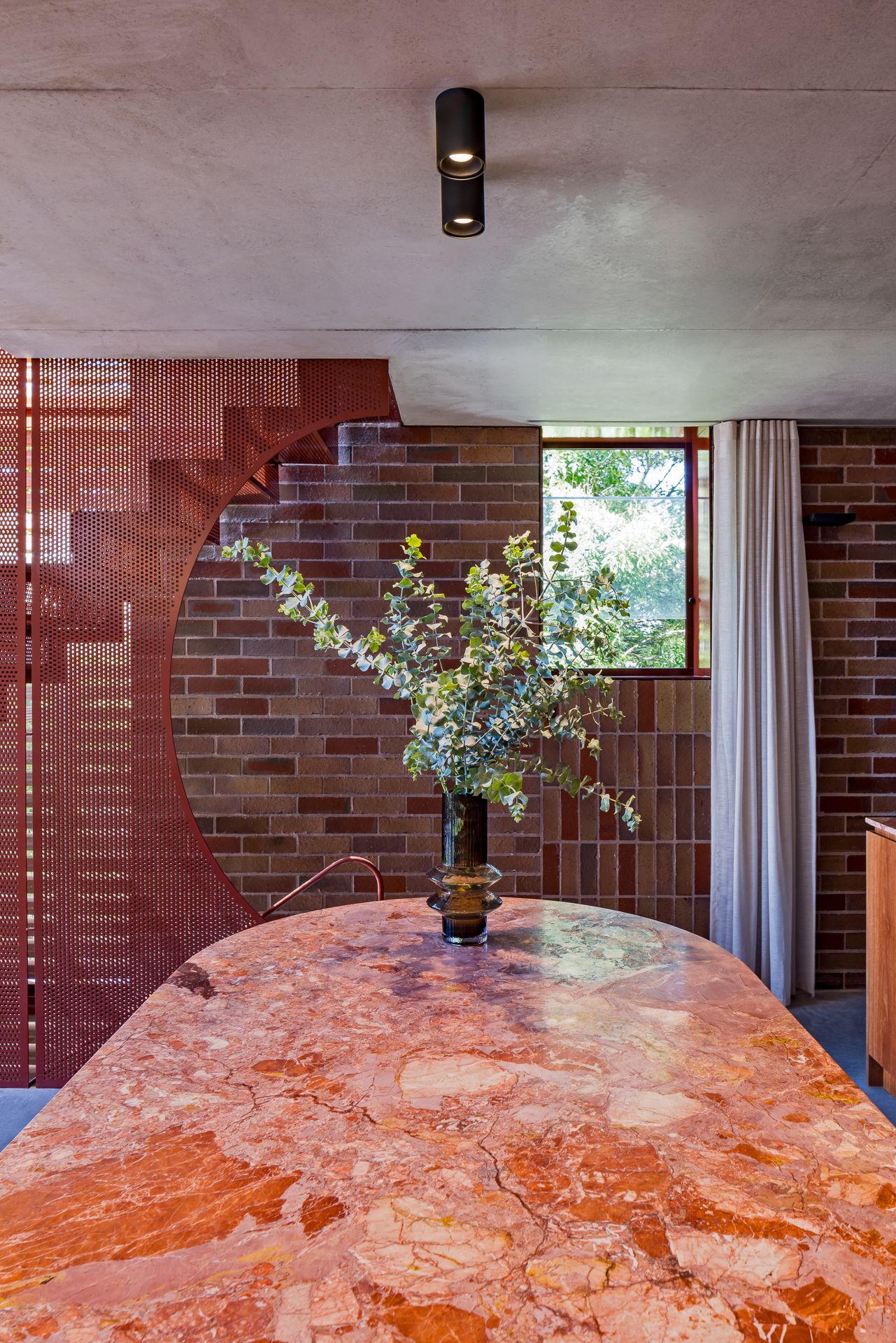 Pink marble table with eucalyptus in a vase beside a curved brick wall, red metal screen and black ceiling spotlights.